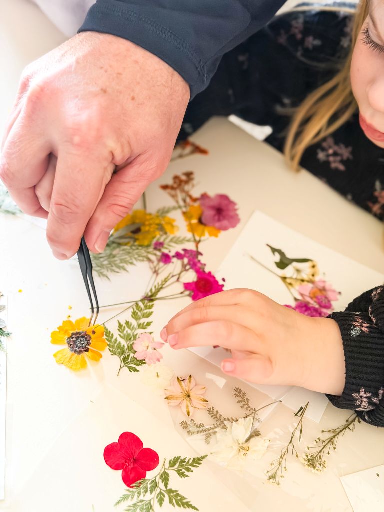 A close-up of a man's hand using crafting tweezers to help a young girl arrange colorful pressed flowers, including yellow zinnias and green ferns, onto a clear sheet for a DIY suncatcher project.