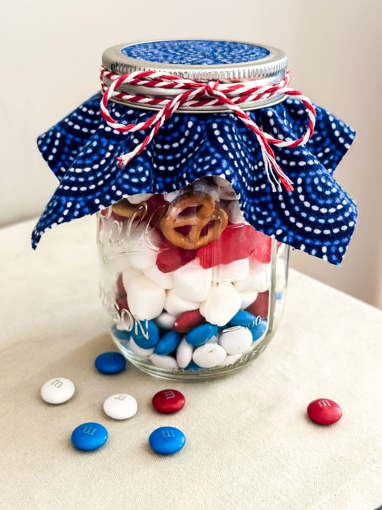 A glass mason jar filled with a patriotic snack mix of red, white, and blue M&M's, marshmallows, and pretzels, topped with blue patterned fabric and tied with red and white baker's twine.