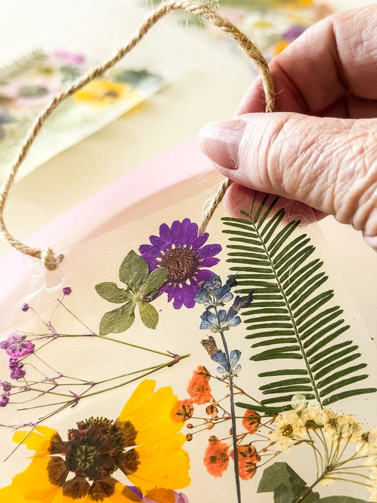 A close-up shot of a hand threading a piece of natural twine through a hole in a DIY suncatcher, which features pressed purple zinnias, yellow wildflowers, green ferns, and delicate baby’s breath.
