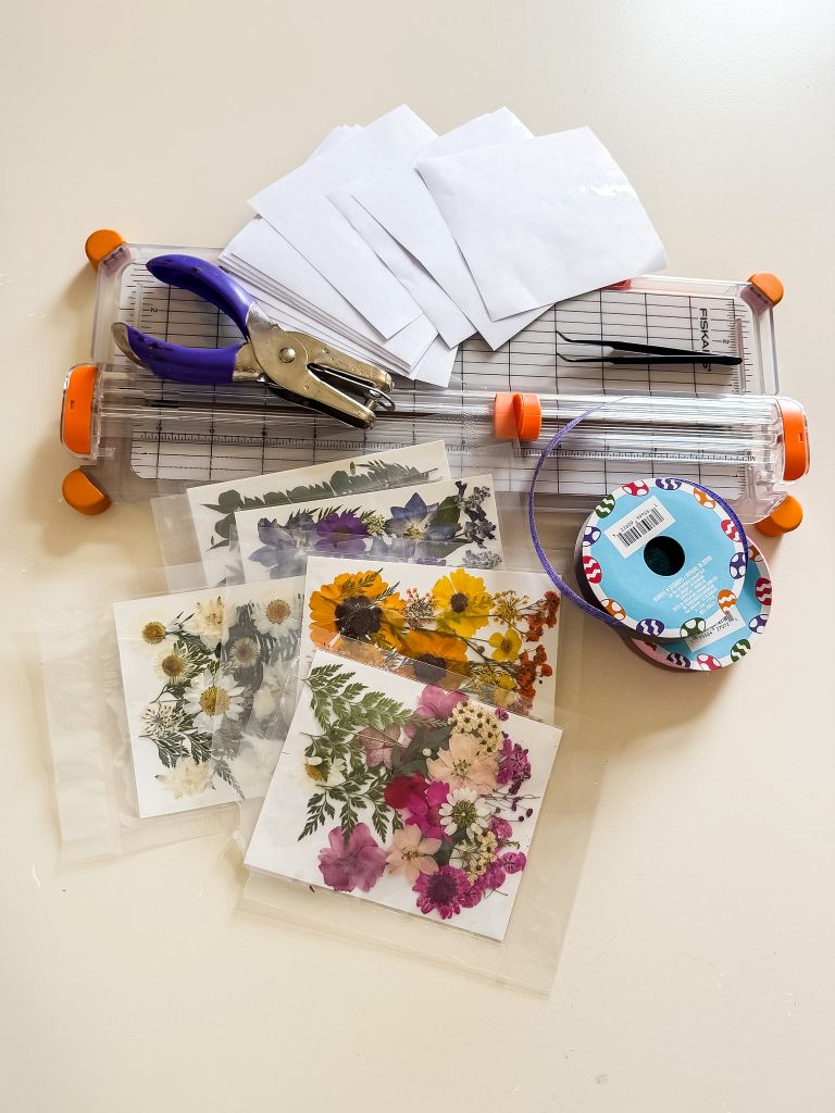 A flat lay of DIY suncatcher supplies including a Fiskars paper trimmer, a hole punch, tweezers, white envelopes, floral patterned ribbon, and several packets of colorful pressed flowers including daisies, ferns, and purple wildflowers.