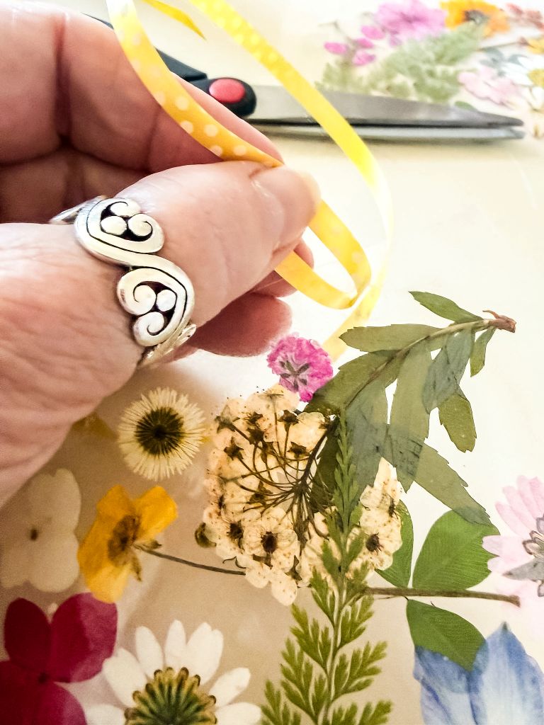 A close-up of a hand with a silver heart-shaped ring holding a bright yellow polka-dot ribbon over a collection of pressed white daisies, yellow wildflowers, and green leaves for a DIY spring suncatcher.