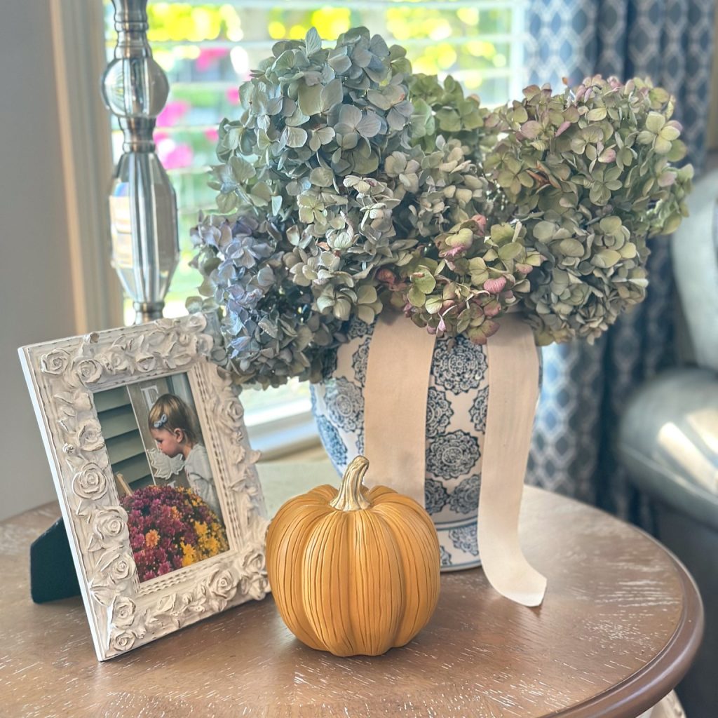 Cottage style vignette featuring dried hydrangeas in a blue and white ginger jar alongside a decorative pumpkin and a vintage floral picture frame.