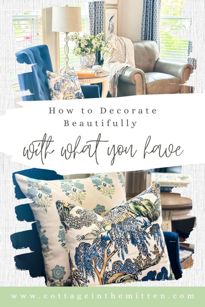 Pinterest pin for Cottage in the Mitten showing a cozy living room with blue accent chairs, patterned throw pillows, a ginger jar vase, and a leather armchair styled with a neutral throw — titled "How to Decorate Beautifully with What You Have".