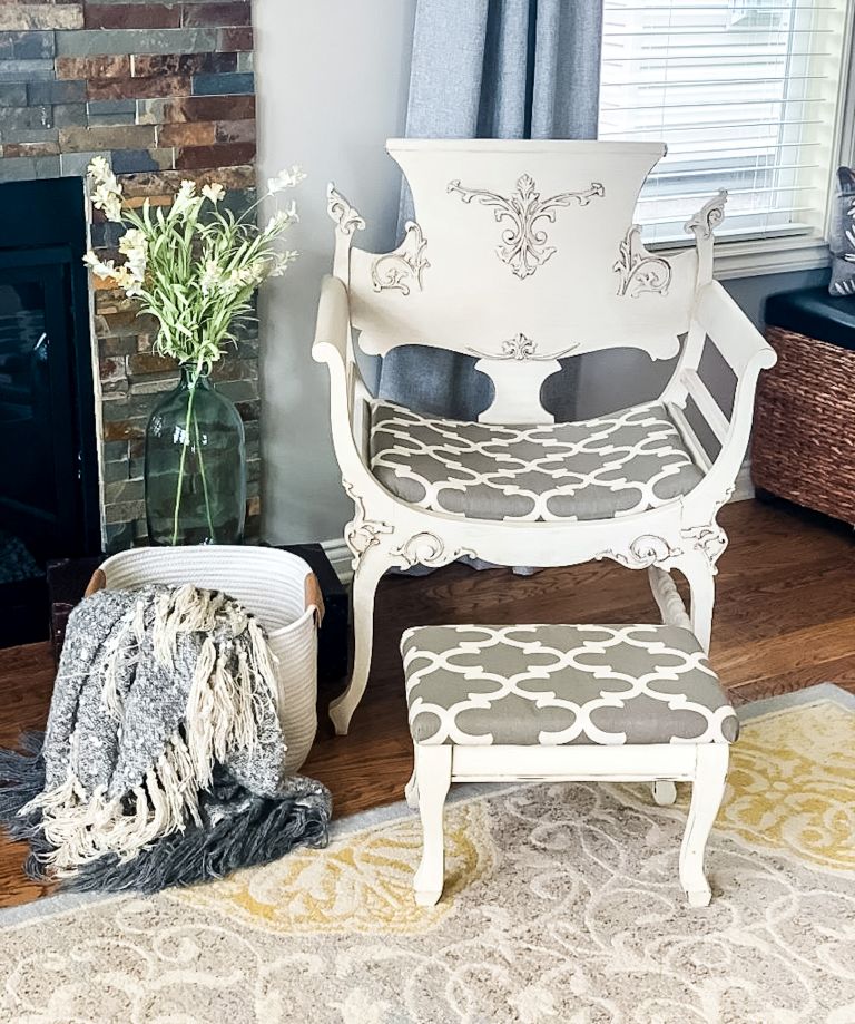 Vintage ornate chair painted white with chalk paint, styled with a matching gray and white geometric upholstered footstool, a cozy gray fringe throw in a rope basket, and fresh stems in a glass vase next to a stone fireplace