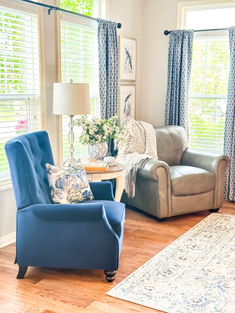 Blue velvet accent chair with chinoiserie throw pillow next to a round side table styled with a blue and white ginger jar, stacked books, and white florals.