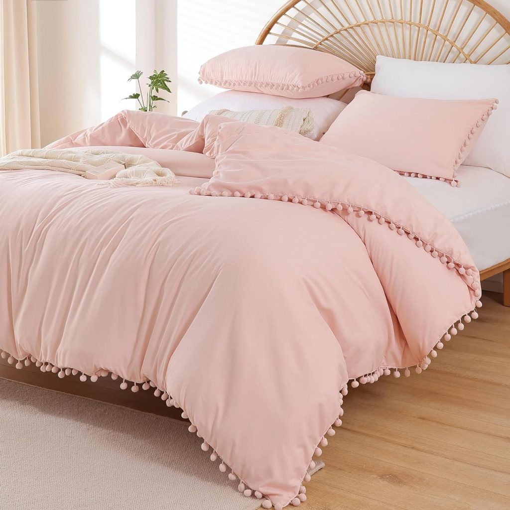 Litanika Blush Pink Boho Pom Pom Comforter Set styled on a cozy bed with matching pink pom-pom pillow shams and a natural curved rattan headboard.