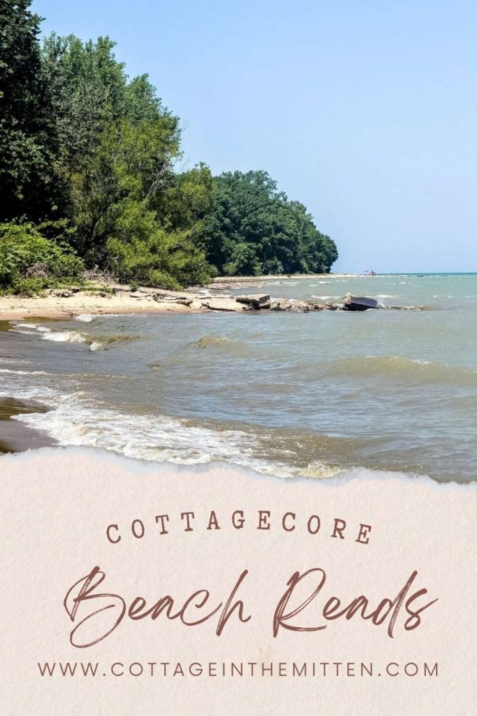 A scenic shoreline with waves lapping against a sandy beach, surrounded by lush green trees under a clear blue sky. Text overlay reads "Cottagecore Beach Reads" with the website URL for Cottage in the Mitten, perfect for summer book recommendations.