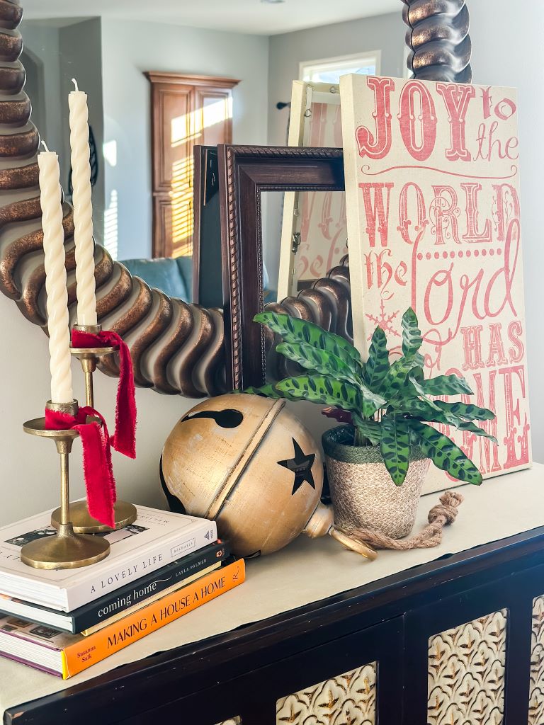 A Christmas tabletop vignette styling idea on a dark sideboard, featuring a stack of vintage books supporting brass candlesticks, a giant gold jingle bell, and a large Christmas canvas sign.