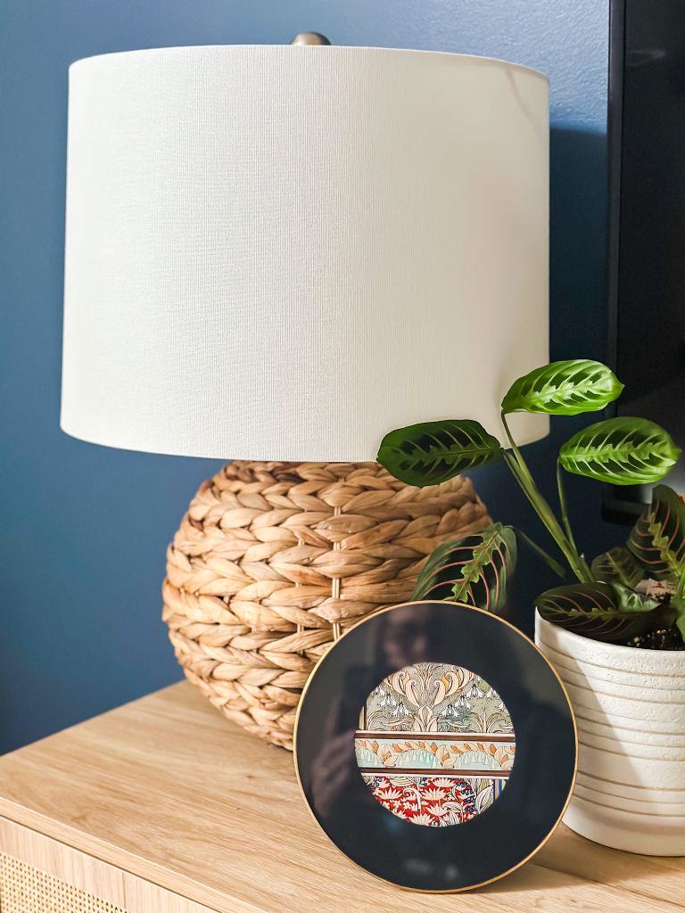 A woven rattan table lamp with a white drum shade styled on a light wood credenza against a dark blue wall, pictured next to a small patterned decorative dish and a green prayer plant in a white textured pot.