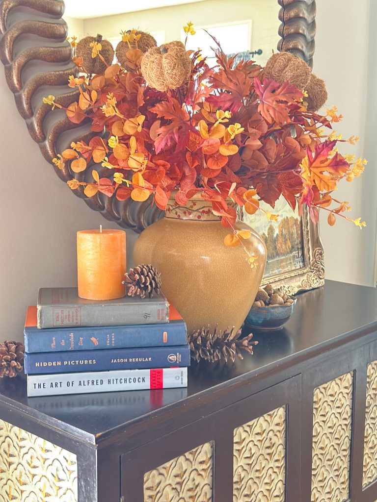 Warm fall tabletop vignette styling on a dark wood sideboard featuring stacked hardcover books, pinecones, and a tall jar of autumn leaves.