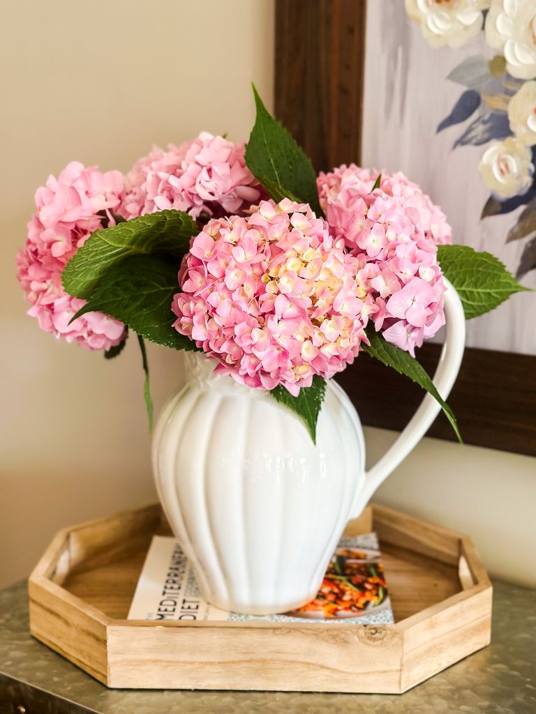 Tabletop vignette styling on a hexagonal wooden tray featuring a white fluted ceramic pitcher with pink hydrangeas.