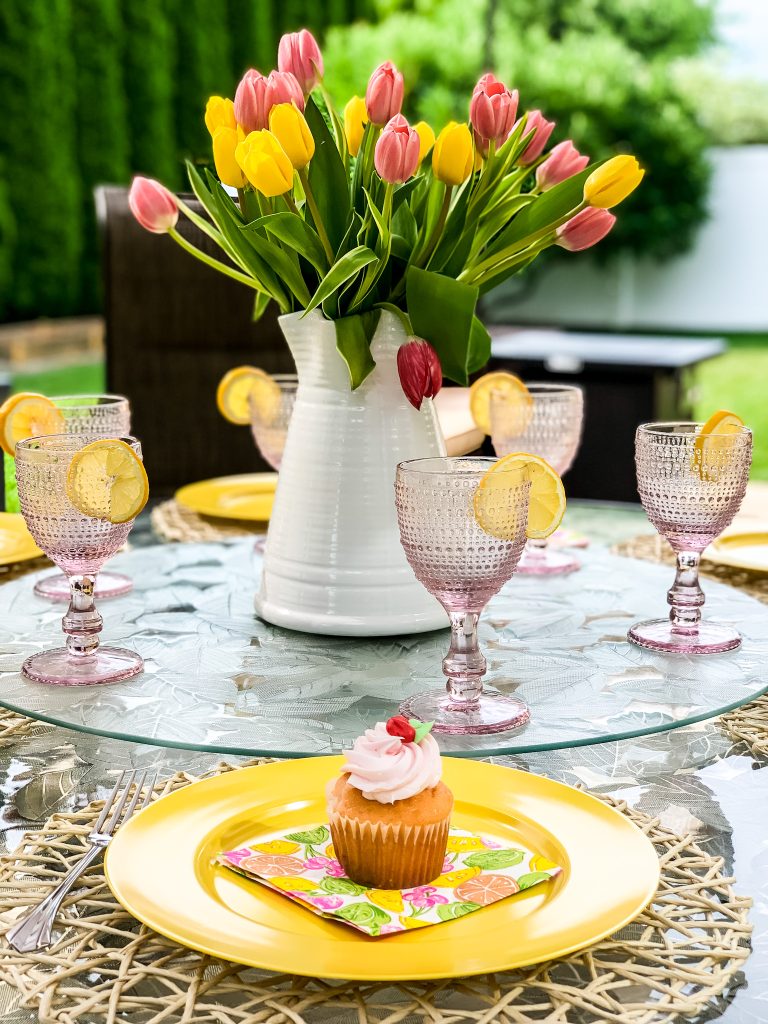 An outdoor dining table styled for a mid summer patio refresh, featuring bright yellow plates, textured pink goblets with lemon slices, a pink frosted cupcake, and a white pitcher filled with a vibrant bouquet of pink and yellow tulips.