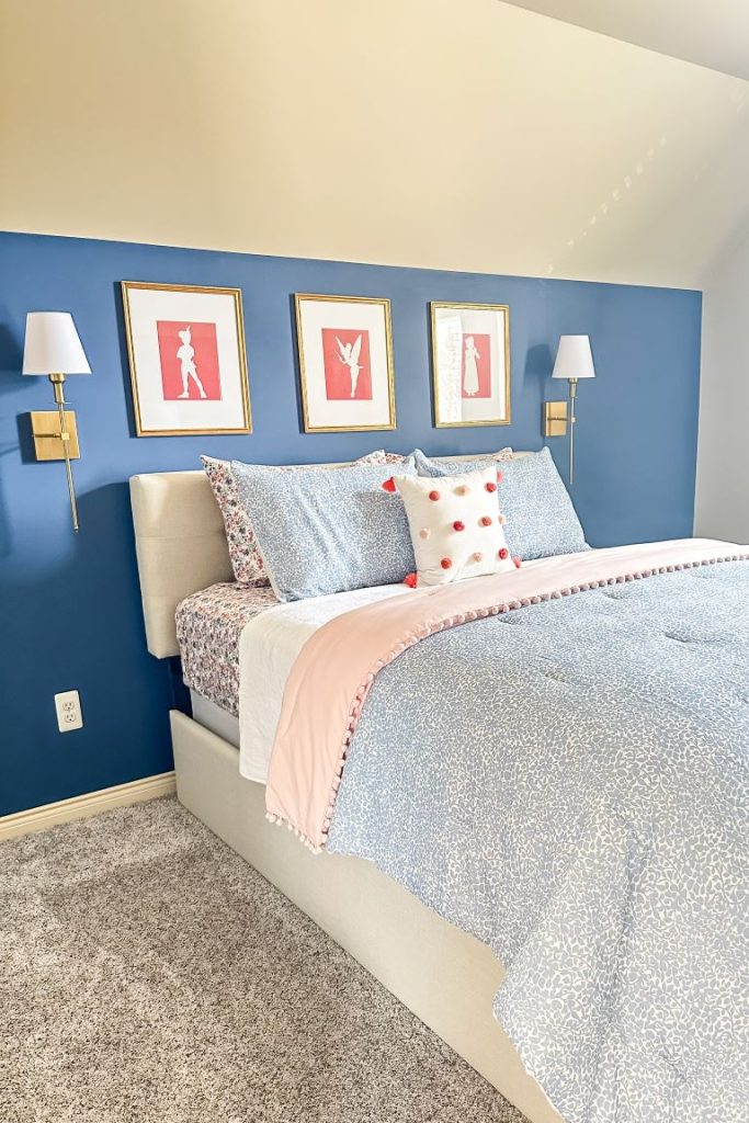 A beautiful guest and granddaughter bedroom featuring a queen bed styled with ditsy floral sheets, a blue patterned coverlet, and a blush pink pom-pom comforter. A beige tufted headboard sits against a denim blue accent knee wall, decorated with three gold-framed pink Peter Pan art prints and gold brass wall sconces.