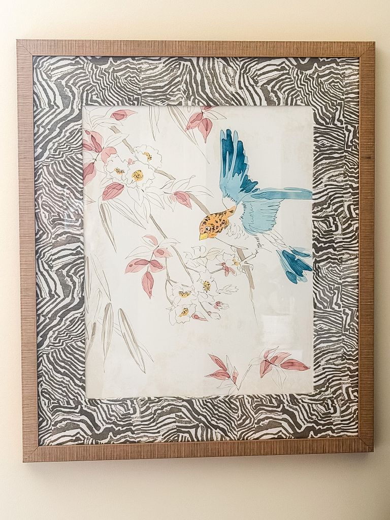 A close-up of a framed watercolor bird art print featuring a blue and yellow bird on a flowering branch, styled with a black and white zebra-patterned mat in a natural wood frame.