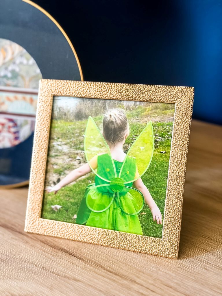 A framed photo of a young girl from behind in a green Tinker Bell fairy costume with wings, styled in a textured gold frame on a wooden credenza for a Peter Pan themed DIY bedroom.