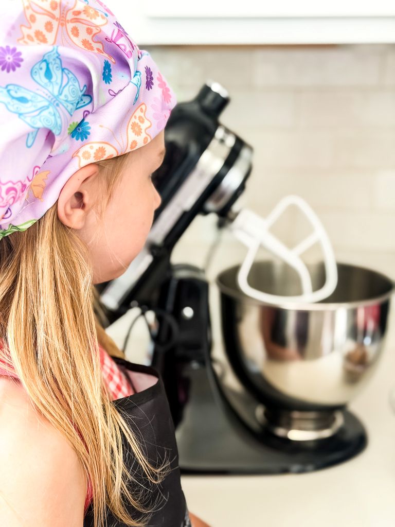 Young girl wearing a butterfly chef hat helping bake fresh peach muffins with a black stand mixer.