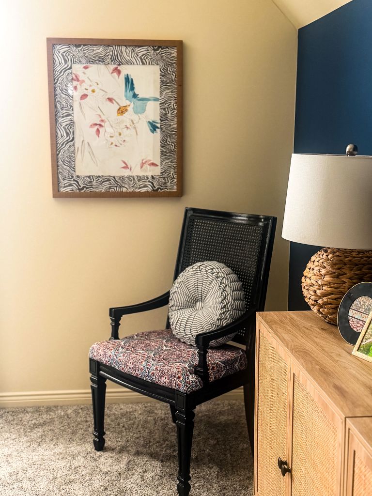 A cozy bedroom corner vignette featuring a vintage-style black painted cane chair with a patterned cushion, styled next to a light wood credenza and a framed bird art print on a cream wall.