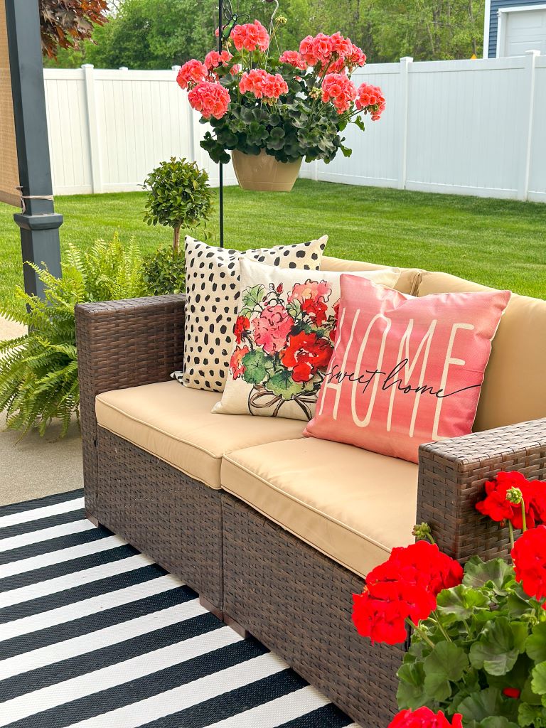 A mid summer patio refresh featuring a brown wicker outdoor sofa styled with coral floral pillows and a black and white striped rug, surrounded by vibrant hanging and potted red geraniums.