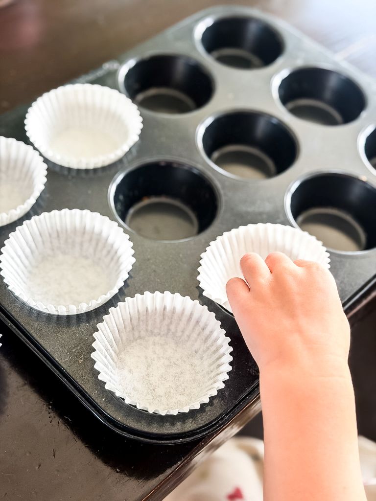 Child's hand placing white paper liners into a metal muffin pan to prepare for baking fresh peach muffins.