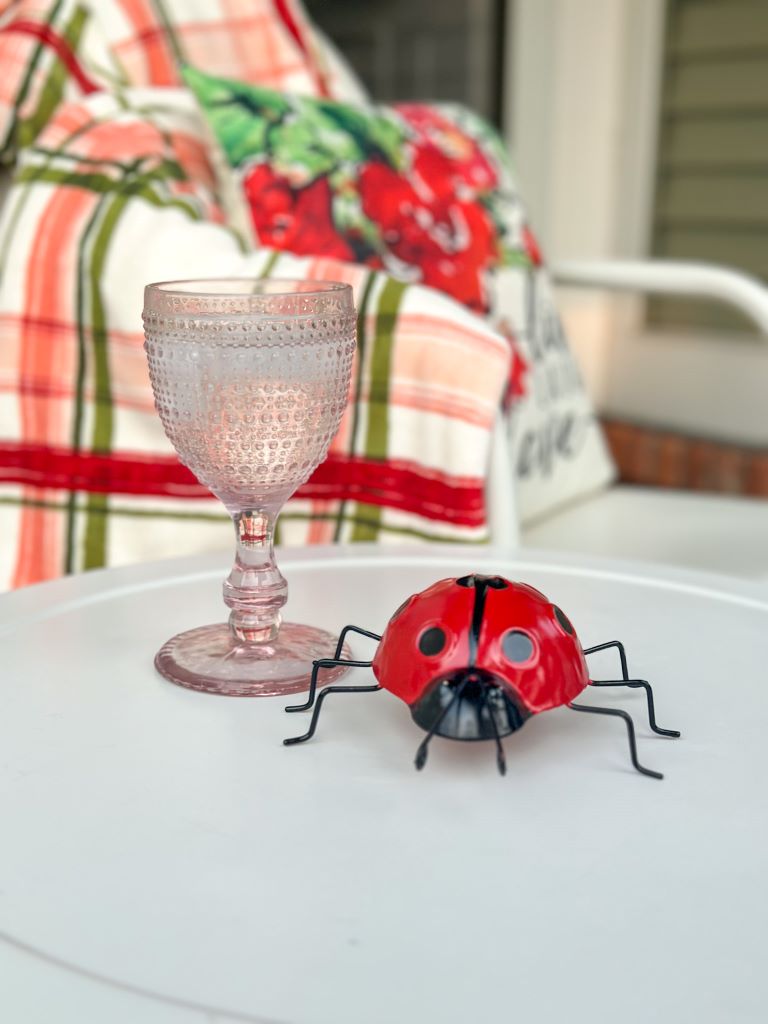 A close-up of a mid summer patio refresh featuring a small red metal ladybug decoration and a pink textured glass goblet on a white side table, with a plaid outdoor cushion in the background.