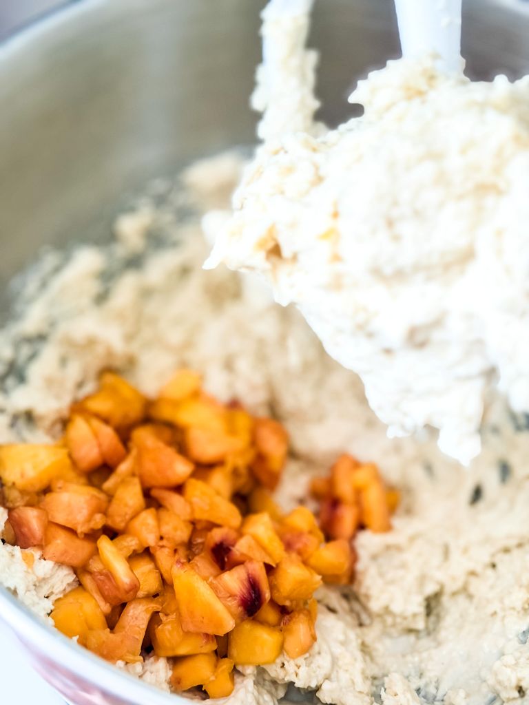 Adding fresh diced peaches into thick muffin batter in a stand mixer bowl.