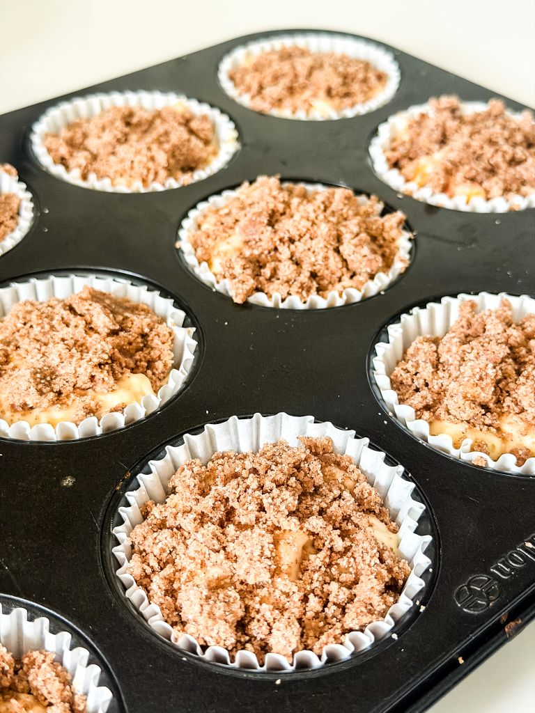Muffin cups filled with raw peach muffin batter and generous cinnamon streusel crumble topping before going into the oven.