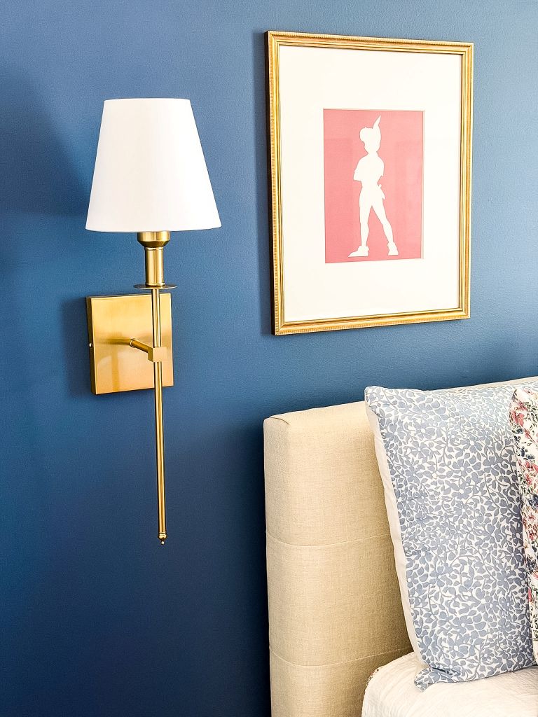A modern brass wall sconce with a white shade mounted next to a beige tufted headboard on a blue wall, featuring a framed pink Peter Pan silhouette and ditsy floral print bedding.