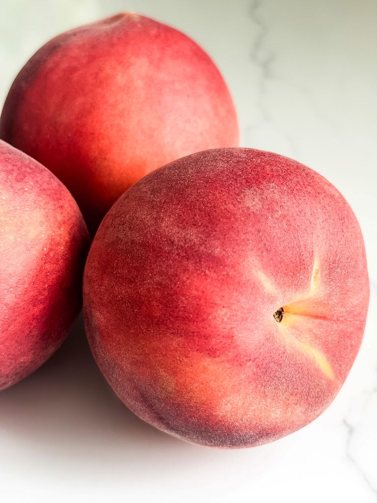Three fresh, ripe peaches resting on a white countertop ready to be chopped for baking peach muffins.