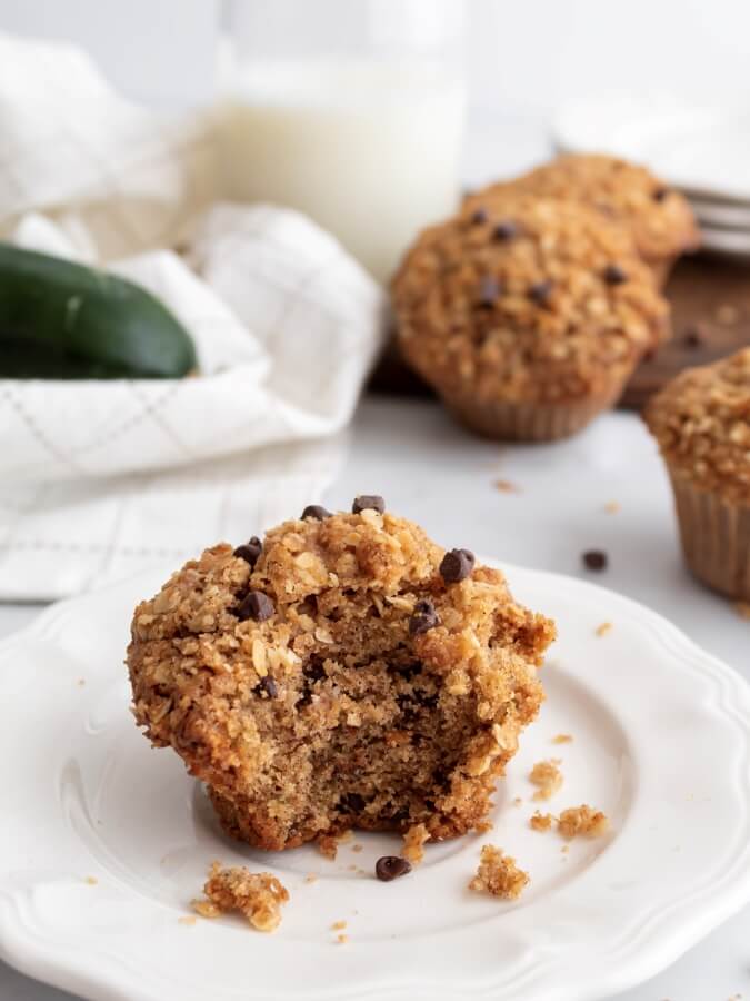 Chocolate chip zucchini muffin with oat streusel topping on a white plate with a bite taken out revealing a tender crumb, with a glass of milk and fresh zucchini in the background.