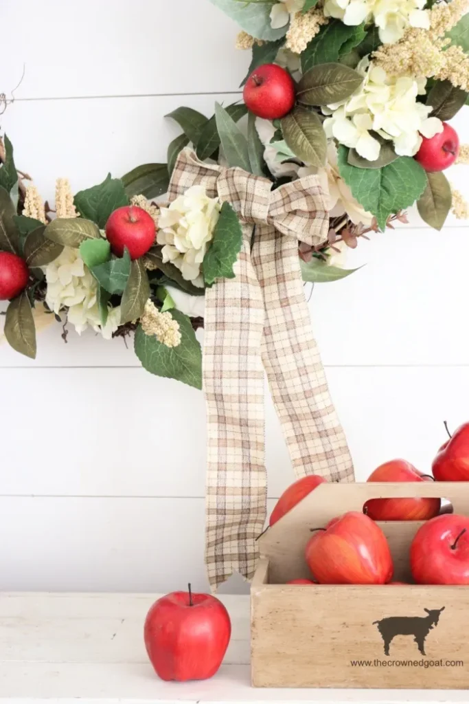 DIY fall grapevine wreath decorated with red apple picks, cream hydrangeas, green leaves, and a plaid bow hanging on a white shiplap wall next to a wooden crate filled with red apples.
