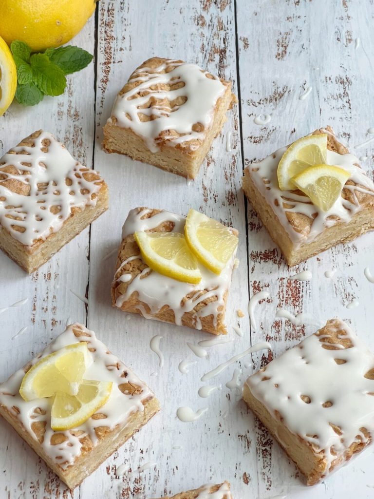Six lemon blondies drizzled with white lemon glaze and topped with fresh lemon slices arranged on a white rustic wood surface with fresh lemons and mint in the background.