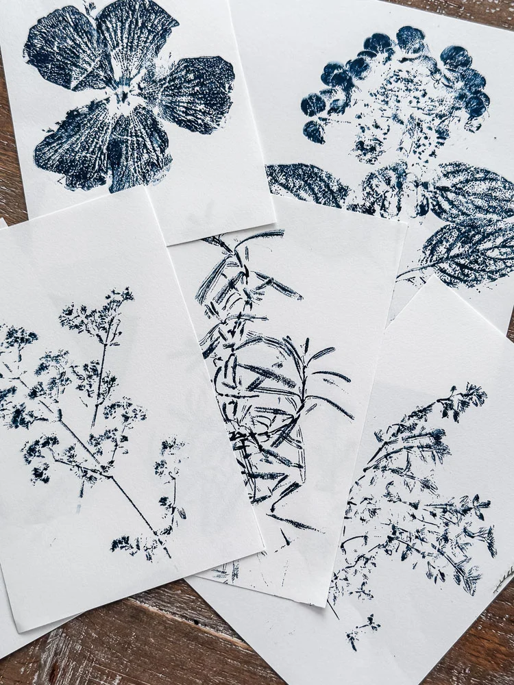 DIY botanical nature prints featuring a pansy, hydrangea, herbs, and wildflowers printed in navy blue ink on white paper spread out on a wooden surface.