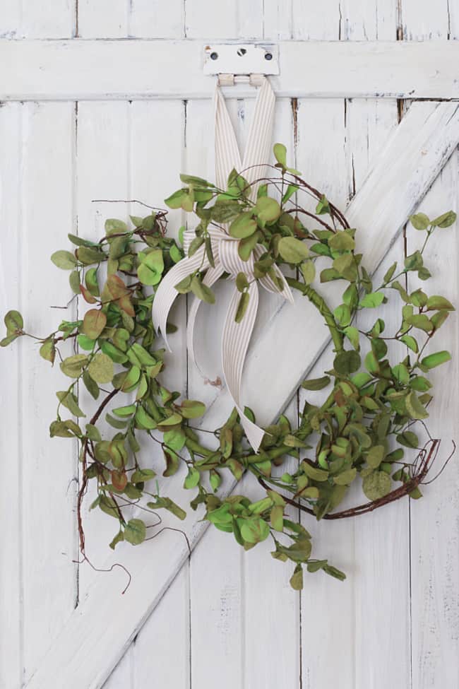 Simple grapevine wreath with green eucalyptus leaves and a cream striped ribbon bow hanging on a white rustic barn door.