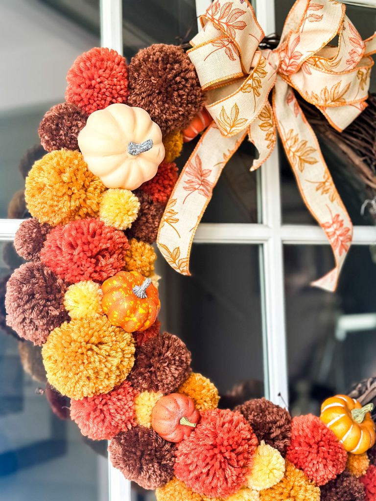 Handmade fall pom-pom wreath in shades of orange, red, yellow, and brown, accented with mini pumpkins and a plaid-and-leaf ribbon bow, hanging on a front door.