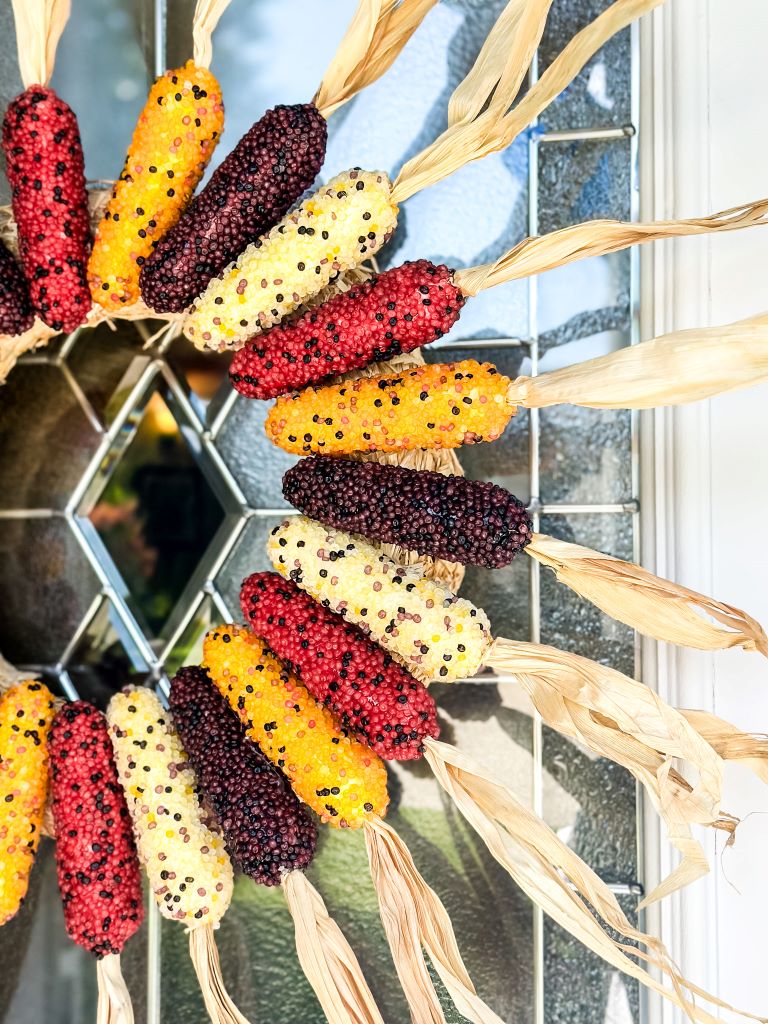 DIY faux Indian corn wreath with multicolor cobs in red, purple, yellow, and cream, arranged with husks radiating outward on a front door.