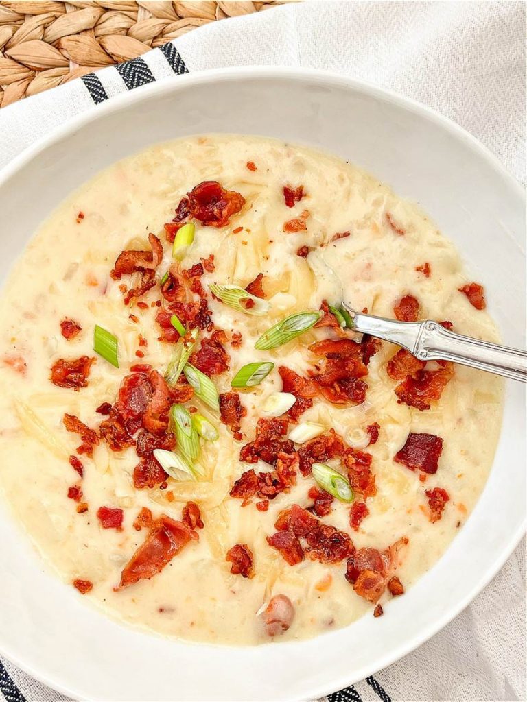 A white ceramic bowl filled with thick, creamy loaded potato soup topped with crispy bacon bits and freshly sliced green onions.