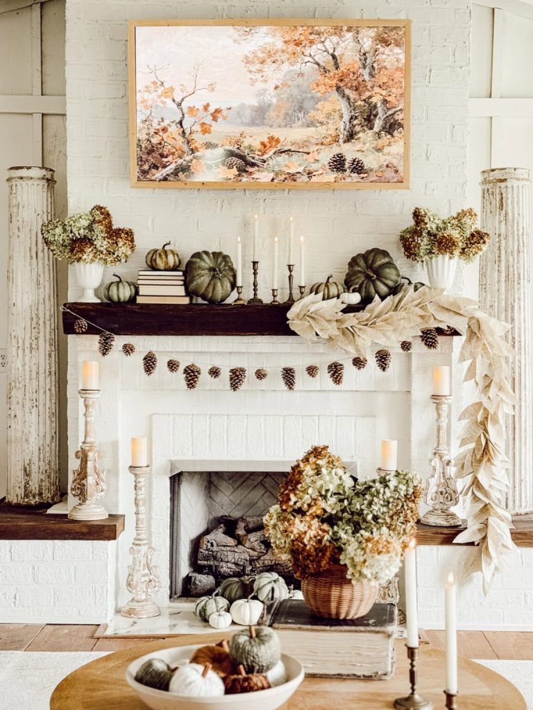Rustic fall fireplace mantel decorated with a simple pinecone garland, dark green heirloom pumpkins, brass candlesticks, dried hydrangeas, and an autumn landscape painting.