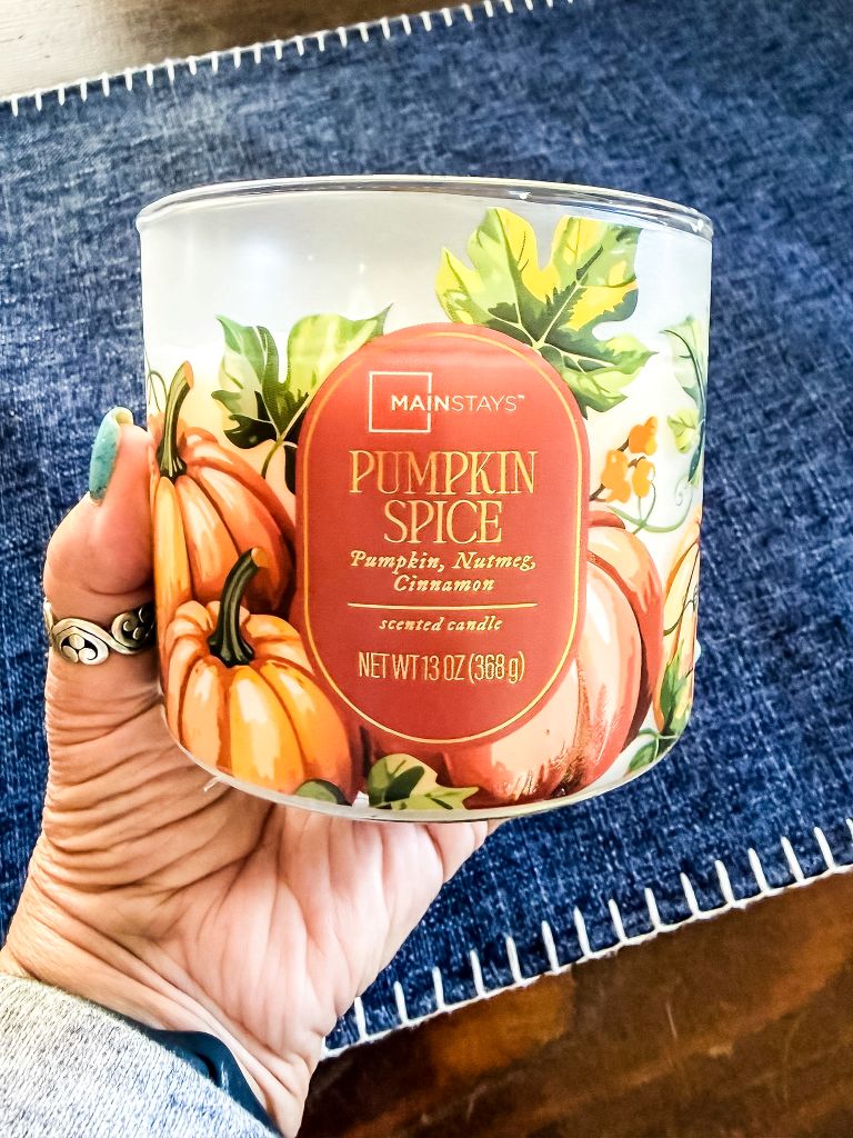 Hand holding a Mainstays Pumpkin Spice scented candle with a pumpkin-print label, set against a denim table runner.
