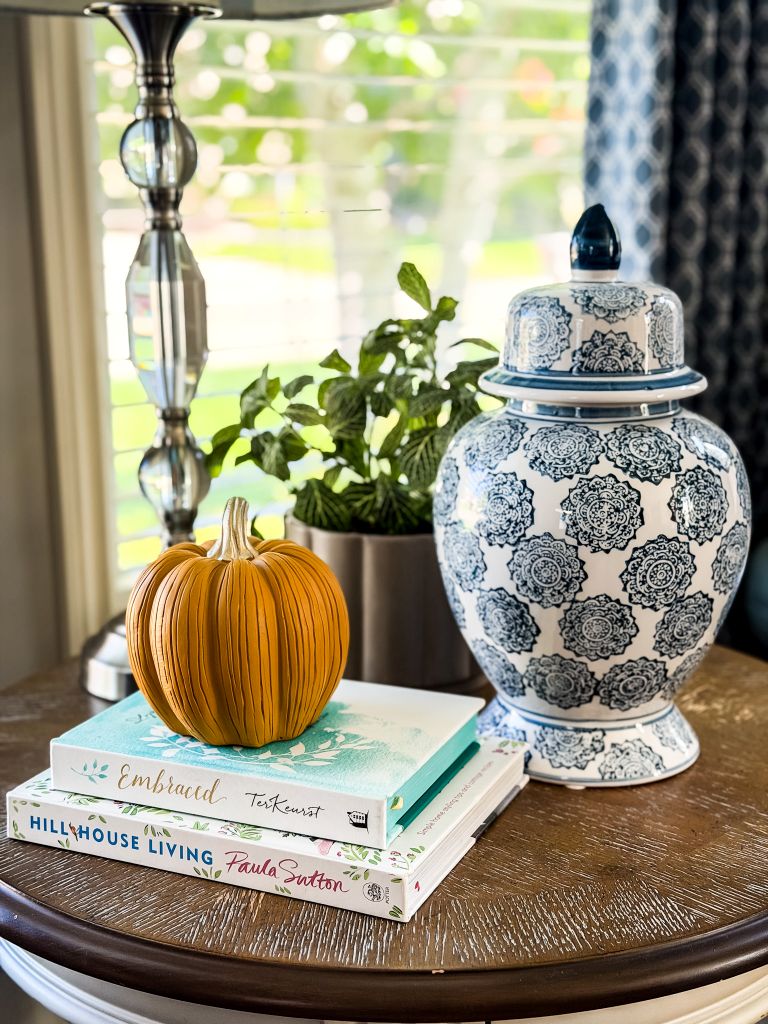 Wood side table styled with a pumpkin on stacked books, a faux greenery plant, and a blue-and-white ginger jar in front of a sunlit window.