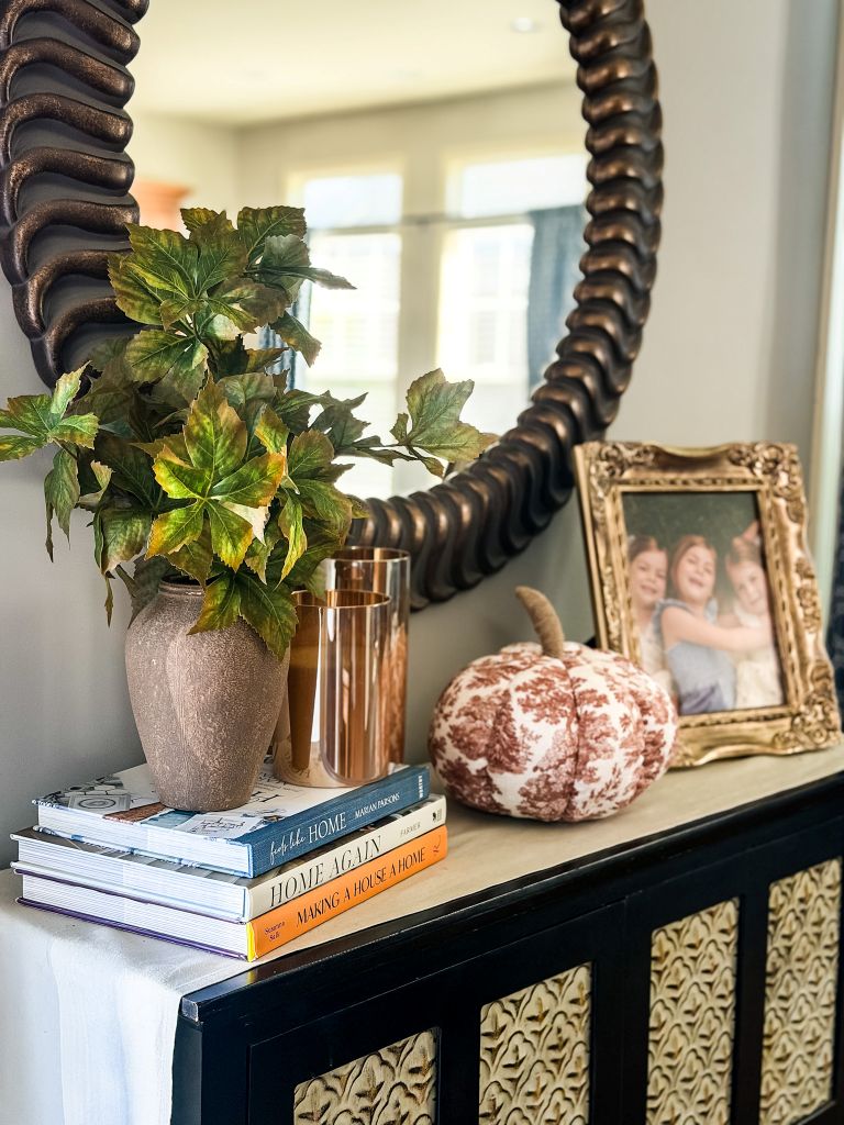 Console table vignette with a vase of faux fall leaves, copper candle holders, a fabric pumpkin, and a framed family photo under a round mirror.